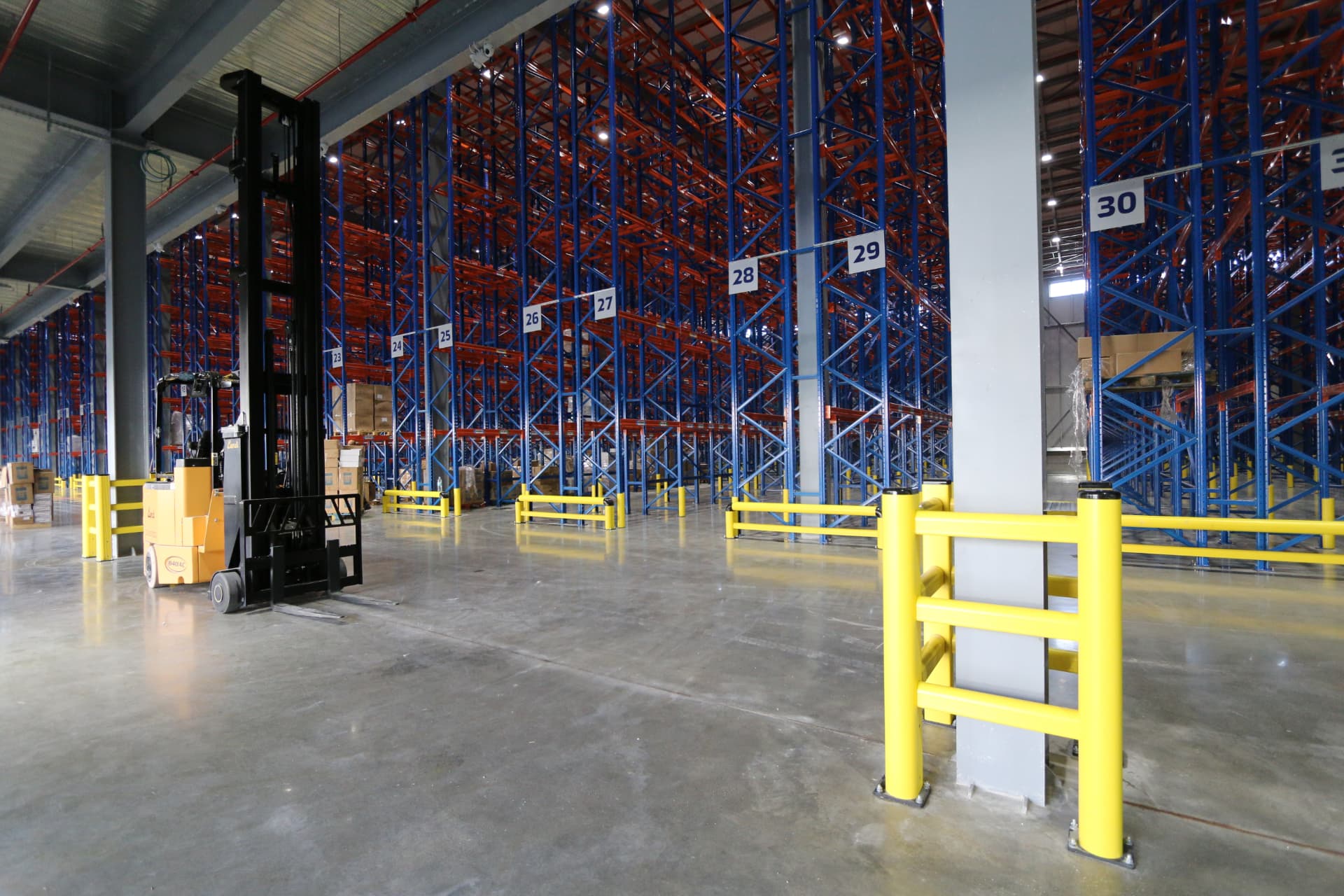 Warehouse aisle with blue and orange pallet racking, yellow safety barriers and a reach truck